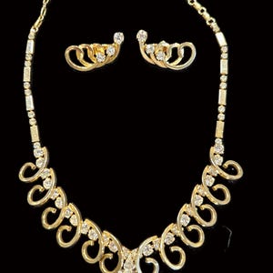 May include: A gold-toned jewelry set featuring a necklace and matching earrings. The necklace has a series of curved, openwork designs adorned with clear gemstones. The earrings mirror the necklace's design. The set is displayed against a black background.