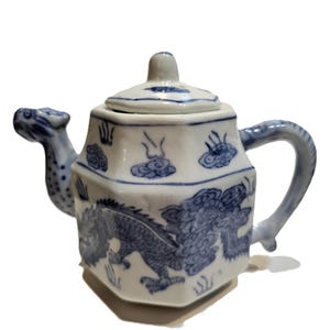 May include: White and blue ceramic teapot with a dragon design. The teapot features a dragon motif in blue, a curved handle, and a spout shaped like a dragon's head. The lid has a small knob and blue cloud patterns.