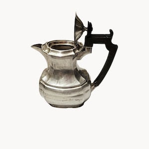 May include: A silver-colored pitcher with a black handle and lid. The pitcher has a bulbous body with horizontal ridges and a spout. The lid is open, revealing the interior. The handle is curved and attached to the side of the pitcher.