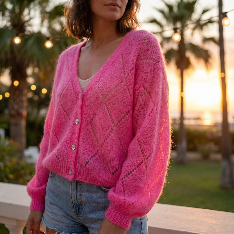 May include: A bright pink, open-weave cardigan with a diamond pattern. The long-sleeved sweater has a V-neck and is buttoned up. The cardigan is paired with denim shorts. The background features palm trees and a sunset.