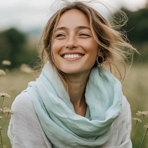 Könnte beinhalten: Eine Frau mit blondem Haar lächelt breit, trägt goldene Ohrringe und einen hellblauen Schal. Der Hintergrund ist ein verschwommenes Feld aus Grün und Gelb mit weichem natürlichem Licht.