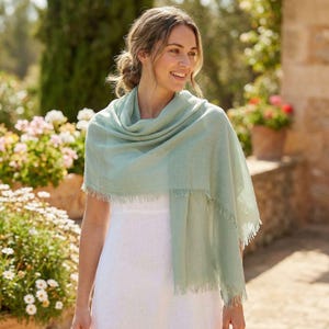Puede incluir: Una bufanda rectangular de color verde salvia claro con una textura tejida suave y bordes con flecos delicadamente deshilachados a lo largo del dobladillo. La bufanda est&aacute; drapeada sobre los hombros, mostrando su tela ligera y tono sutil.
