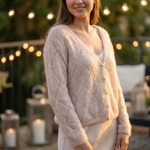 May include: A light beige cardigan with a diamond pattern and button closure. The V-neck sweater is made of a soft, textured knit fabric. The cardigan is a neutral color and has a relaxed fit.