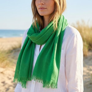 Puede incluir: Una bufanda de color verde brillante con una textura suave y ligera, con bordes deshilachados, sobre una camisa blanca de botones. La bufanda presenta un tono esmeralda s&oacute;lido y vibrante, con una forma rectangular que cae sobre el cuello y hombros.