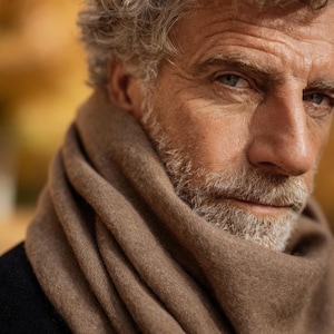 Può includere: Ritratto in primo piano di una persona con capelli grigi e barba folta, che indossa una morbida sciarpa marrone chiaro. La sciarpa è drappeggiata attorno al collo e alle spalle, creando un aspetto caldo e invitante. Lo sfondo è sfocato, con toni caldi.