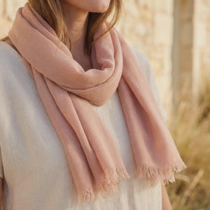 Puede incluir: Una bufanda de color rosa claro suave con una apariencia tejida y texturizada, con bordes delicadamente deshilachados en la parte inferior. La bufanda est&aacute; drapeada holgadamente alrededor del cuello, con un color s&oacute;lido uniforme y tejido visible.
