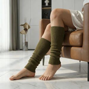 Könnte beinhalten: Olivgrüne Beinwärmer aus Rippstrick. Diese Wärmer sind so konzipiert, dass sie vom Knöchel bis zum Knie getragen werden können. Die Beinwärmer bestehen aus einem weichen, dehnbaren Material. Die Person auf dem Bild sitzt auf einem braunen Ledersessel.