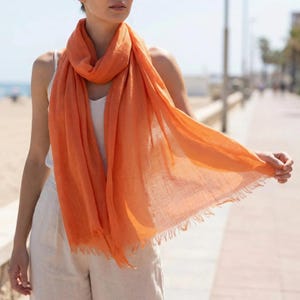 Puede incluir: Una bufanda de color naranja brillante con una textura ligera, semitransparente y bordes deshilachados. La bufanda est&aacute; drapeada alrededor del cuello y los hombros, con un tejido suelto y una apariencia suave y fluida que cuelga hacia un lado.