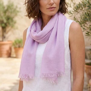 Puede incluir: Una bufanda de color lavanda claro con una textura tejida suave y bordes deshilachados, colocada alrededor del cuello sobre una sencilla camiseta blanca sin mangas. La bufanda tiene forma rectangular y una tela ligera que cuelga suelta.
