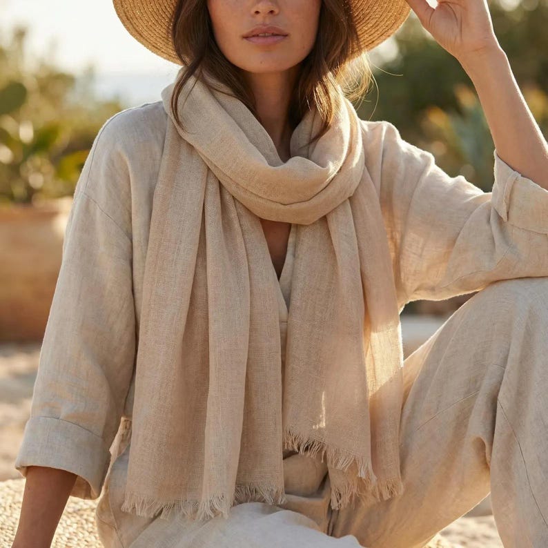 Puede incluir: Una bufanda de color beige con tejido texturizado y bordes deshilachados, colocada alrededor del cuello. La bufanda se combina con una camisa beige de manga larga y un sombrero de paja, creando un conjunto coordinado en tonos beige claro.