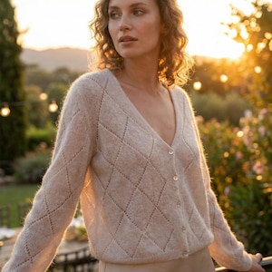 Könnte beinhalten: Hellbeige Cardigan mit Rautenmuster und V-Ausschnitt. Der langärmlige Pullover hat drei Knöpfe vorne. Der Cardigan ist aus einem weichen, strukturierten Material gefertigt.