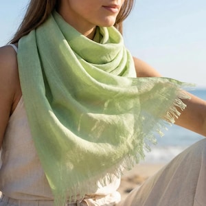 Puede incluir: Una bufanda de color verde claro con un tejido suave y texturizado y delicados bordes deshilachados. La tela est&aacute; drapeada holgadamente, mostrando una calidad semitransparente y un aspecto natural y ligero. La bufanda presenta flecos en los bordes.