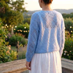 May include: A light blue knit sweater with a diamond pattern. The sweater has long sleeves and a crew neck. The person is wearing a white skirt. The background includes a wooden bench, greenery, and soft lighting.