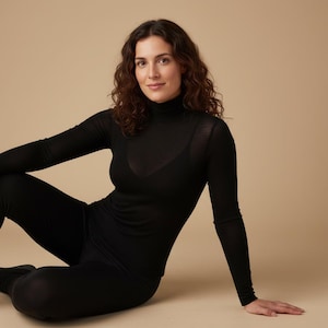 Può includere: Completo nero composto da dolcevita a maniche lunghe e leggings abbinati. L'outfit è realizzato in un materiale sottile e aderente. La modella è seduta con le gambe incrociate, su uno sfondo beige.