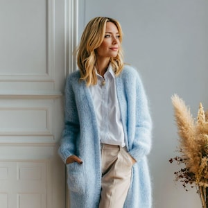 Peut inclure: Un long cardigan bleu clair à devant ouvert avec des poches. Le cardigan est porté sur une chemise blanche à col et un pantalon beige. La femme se tient près d'une porte blanche et d'un vase de fleurs séchées.