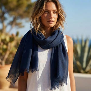 Puede incluir: Una bufanda de color azul marino s&oacute;lido con una tela ligera y texturizada con bordes deshilachados. La bufanda est&aacute; colocada holgadamente alrededor del cuello, presentando una forma rectangular con hilos tejidos visibles y un acabado mate.