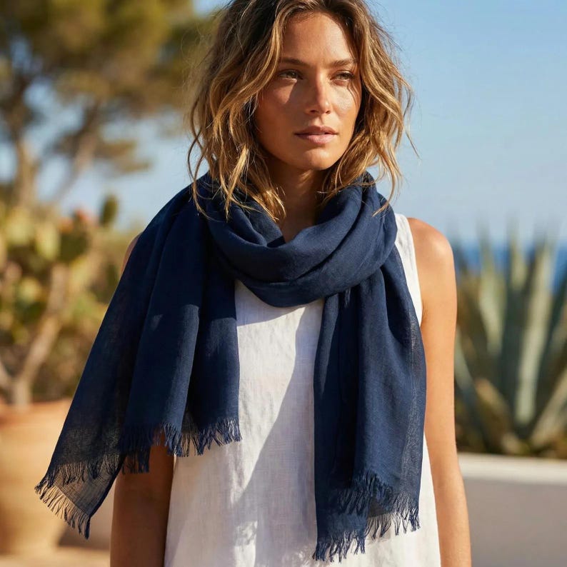 Puede incluir: Una bufanda de color azul marino s&oacute;lido con una tela ligera y texturizada con bordes deshilachados. La bufanda est&aacute; colocada holgadamente alrededor del cuello, presentando una forma rectangular con hilos tejidos visibles y un acabado mate.