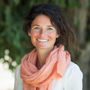 May include: A woman with brown hair and a peach-coloured scarf smiles at the camera. She is wearing a white shirt. The background is blurred green foliage.