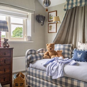 May include: A cozy child's bedroom with a blue and white gingham patterned daybed, teddy bears, and a blue striped shirt. The room features a wooden dresser, a wicker house-shaped basket, and a decorative elephant head on the wall.