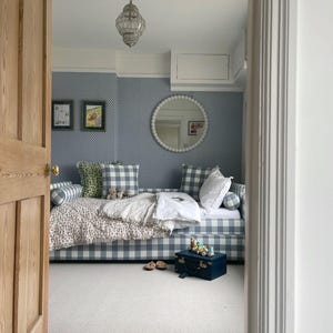 May include: Interior shot of a bedroom with a daybed upholstered in blue and white gingham fabric. The room includes blue patterned wallpaper, a round mirror, and a small blue trunk. A decorative chandelier hangs from the ceiling. The room has a light, airy feel.