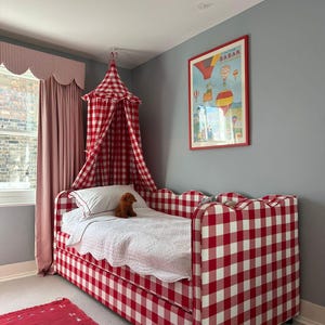 Op de afbeelding: Een dagbed bekleed met rood-witte vichy-ruitstof met een geschulpt hoofd- en voeteneinde, gecombineerd met een bijpassende rood-witte hemel, een witte doorgestikte sprei en een kleine bruine knuffel op het bed.