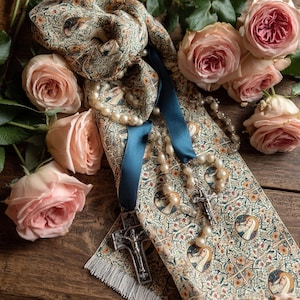May include: A detailed shot of a patterned scarf with a blue ribbon and a pearl rosary. A silver crucifix hangs from the scarf. Soft pink roses are arranged around the scarf on a wooden surface.