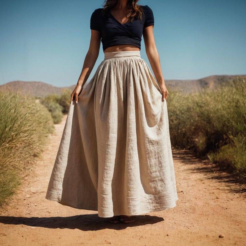 May include: A woman wearing a black crop top and a long, flowing beige linen skirt. The skirt has a high waist and is gathered at the front. She is standing on a dirt path with tall grass on either side.