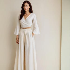 May include: A cream-colored linen two-piece outfit. The top is a wrap-style blouse with long, flared sleeves. The high-waisted pants have a wide leg and pockets. The outfit is made of a lightweight fabric.