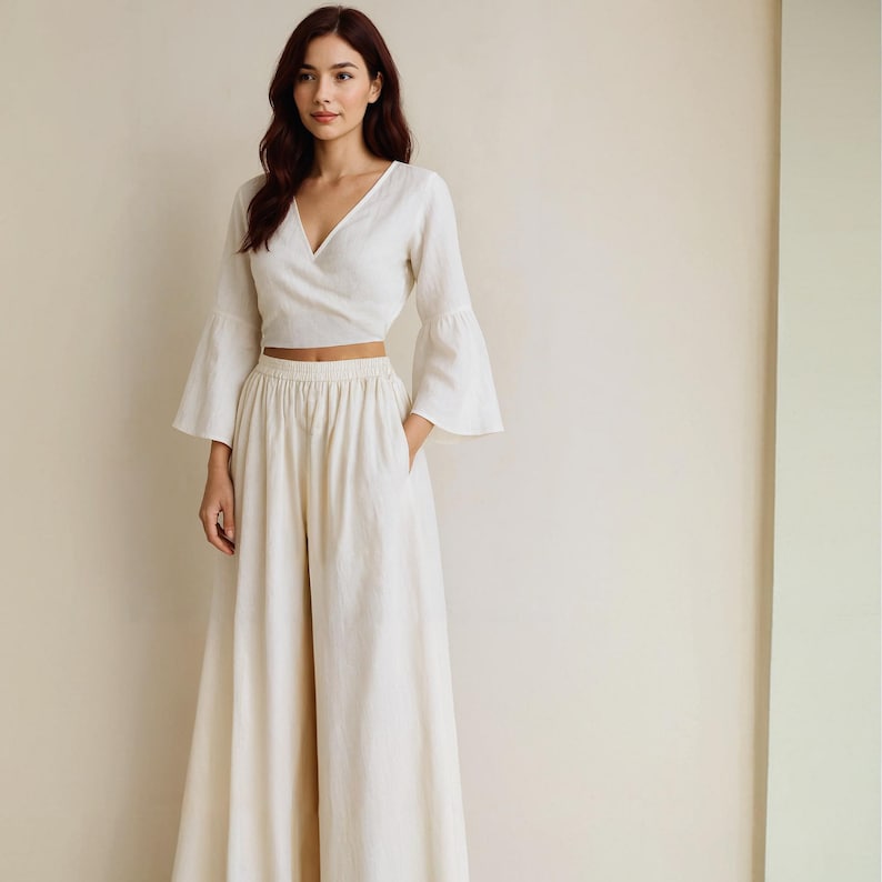 May include: A cream-colored linen two-piece outfit. The top is a wrap-style blouse with long, flared sleeves. The high-waisted pants have a wide leg and pockets. The outfit is made of a lightweight fabric.
