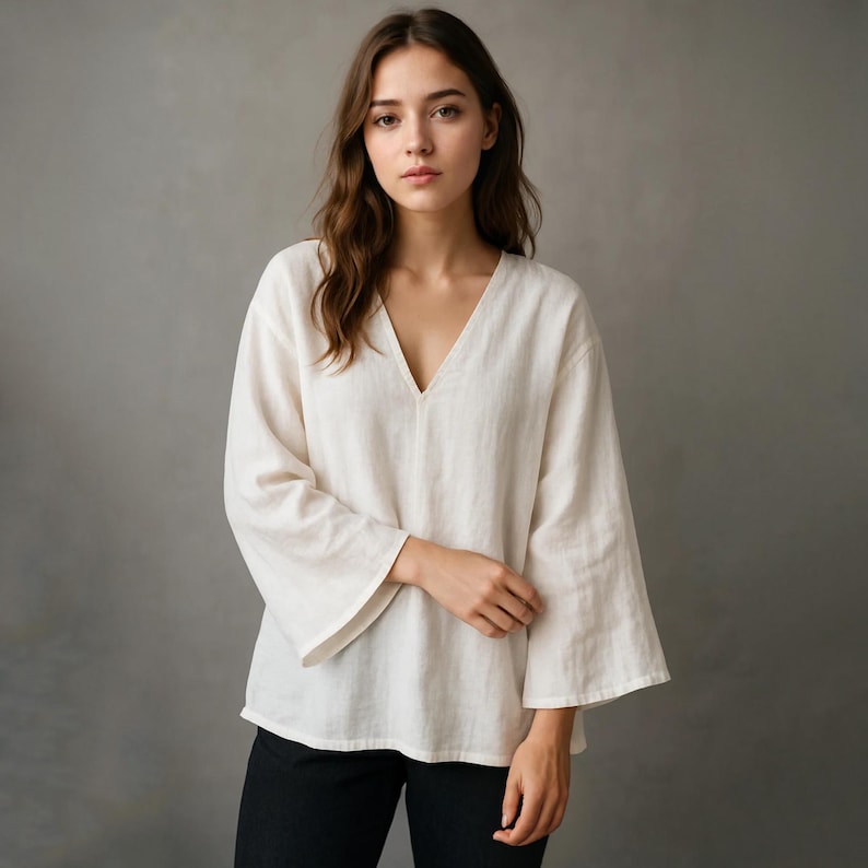 Op de afbeelding: Een cr&egrave;mekleurige linnen blouse met een diepe V-hals en wijde, uitlopende mouwen. De top met lange mouwen is gemaakt van een lichte stof en wordt gecombineerd met een zwarte broek. Het model staat voor een grijze achtergrond.