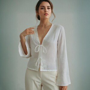 May include: A white linen blouse with a deep V-neck and long, flared sleeves. The blouse has tie closures at the neckline and waist. The model is wearing the blouse with cream-coloured trousers.