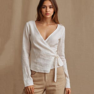 May include: A white wrap top with long sleeves and a V-neck. The top is tied at the waist with a matching fabric tie. The fabric appears to be a lightweight linen or similar material. The model is wearing tan pants.