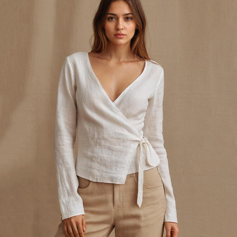 May include: A white wrap top with long sleeves and a V-neck. The top is tied at the waist with a matching fabric tie. The fabric appears to be a lightweight linen or similar material. The model is wearing tan pants.