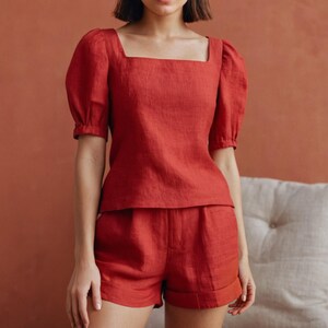 以下が含まれることがあります： 赤いリネンのツーピースセット。トップスはスクエアネックで、短いパフスリーブが付いています。お揃いのショートパンツはハイウエストで、裾はロールアップされています。軽量の生地は、夏の服装を示唆しています。