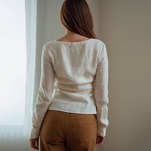 May include: A cream-colored long-sleeve shirt with a scoop neckline. The shirt is paired with brown pants. The model is facing away from the camera, standing in front of a window with white curtains.