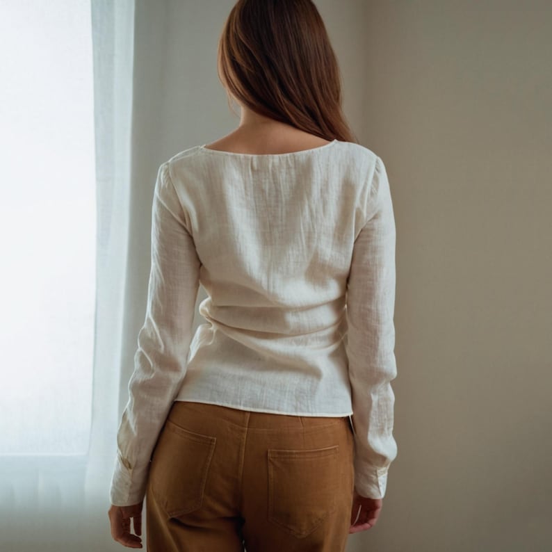May include: A cream-colored long-sleeve shirt with a scoop neckline. The shirt is paired with brown pants. The model is facing away from the camera, standing in front of a window with white curtains.