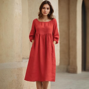 May include: A red linen dress with a round neckline and three-quarter sleeves. The dress has a gathered waist and pockets. The model is standing in front of a stone wall.