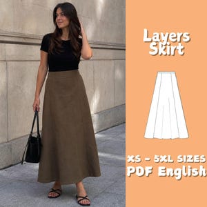 May include: A long olive green skirt with a high waist and a flared silhouette. The image includes a white line drawing of the skirt pattern labeled Layers Skirt, with text indicating XS - 5XL SIZES and PDF English for the sewing pattern.