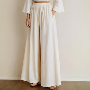 May include: Cream-colored wide-leg pants with an elastic waistband and pockets. The pants are paired with a matching crop top with flared sleeves. The model is wearing gold sandals. The outfit is made of a lightweight fabric.