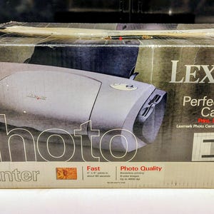 May include: A rectangular cardboard box for a Lexmark P707 photo printer. The box is primarily dark gray and white, featuring an image of the silver printer, the Lexmark logo, and text including "Perfect for Digital Camera Users" and "Photo Printer."