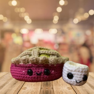 May include: A crocheted pie and a dumpling, both with smiling faces. The pie has a pink base, a tan crust, and a green filling. The dumpling is white. Both have black button eyes and stitched smiles.