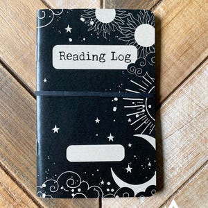 May include: Black reading log notebook with white celestial illustrations, including stars, suns, and moons. The words "Reading Log" are printed on a white label. A black elastic band secures the notebook. The design is labeled "DESIGN II".