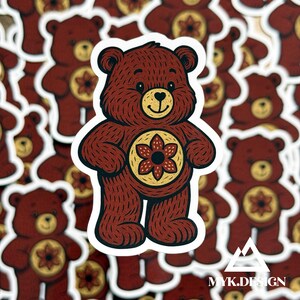 May include: A die-cut sticker featuring a brown bear with a textured fur pattern, a cream-colored muzzle, and a circular emblem on its chest containing a dark red six-petaled flower design. The sticker has a white border and a matte finish.
