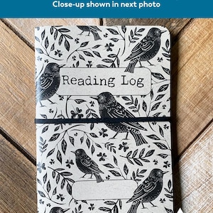 May include: A Reading Log notebook with a black and white bird and leaf pattern. The cover features the text "Reading Log" and is held closed by a black elastic band. The notebook has a minor corner crease from manufacturing.