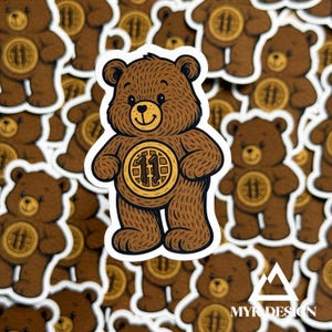 May include: A brown bear sticker with a white border featuring a waffle-shaped emblem on its chest with the number 11 printed in the center. The bear has a smiling face, dark brown fur texture, and rounded ears, presented as a die-cut sticker design.