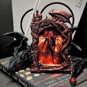 May include: A detailed, reddish-brown dragon-themed picture frame with a castle design. The frame holds an image of a figure with wings, set against a fiery background. Black dragon sculptures and books with the title "That Other World" are also visible.