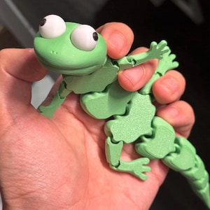 Peut inclure: Un jouet en forme de grenouille verte articulée, avec de grands yeux blancs et des pupilles noires. La grenouille est tenue dans une main, mettant en valeur ses membres mobiles et son corps segmenté. Le jouet est en matériau vert clair.