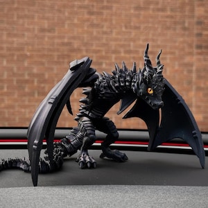 May include: A black dragon figurine with intricate detailing, including large wings and a spiky back. The dragon has yellow eyes and is positioned on a dark surface. The background features a brick wall. A fantasy-themed collectible.