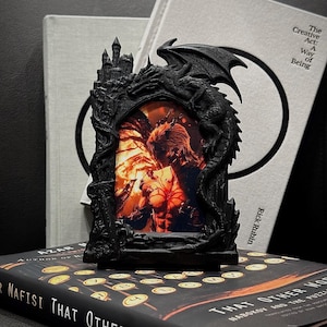 Puede incluir: Marco de fotos gótico negro con diseño de dragón y castillo. El marco contiene una imagen colorida de una persona. El marco está sobre un libro titulado "THAT OTHER WOR". El nombre del autor también es visible.