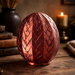 May include: A decorative, metallic red egg-shaped object with a textured, geometric pattern. The egg is sitting on a wooden surface. The background includes a candle, books, and a vase with dried plants.